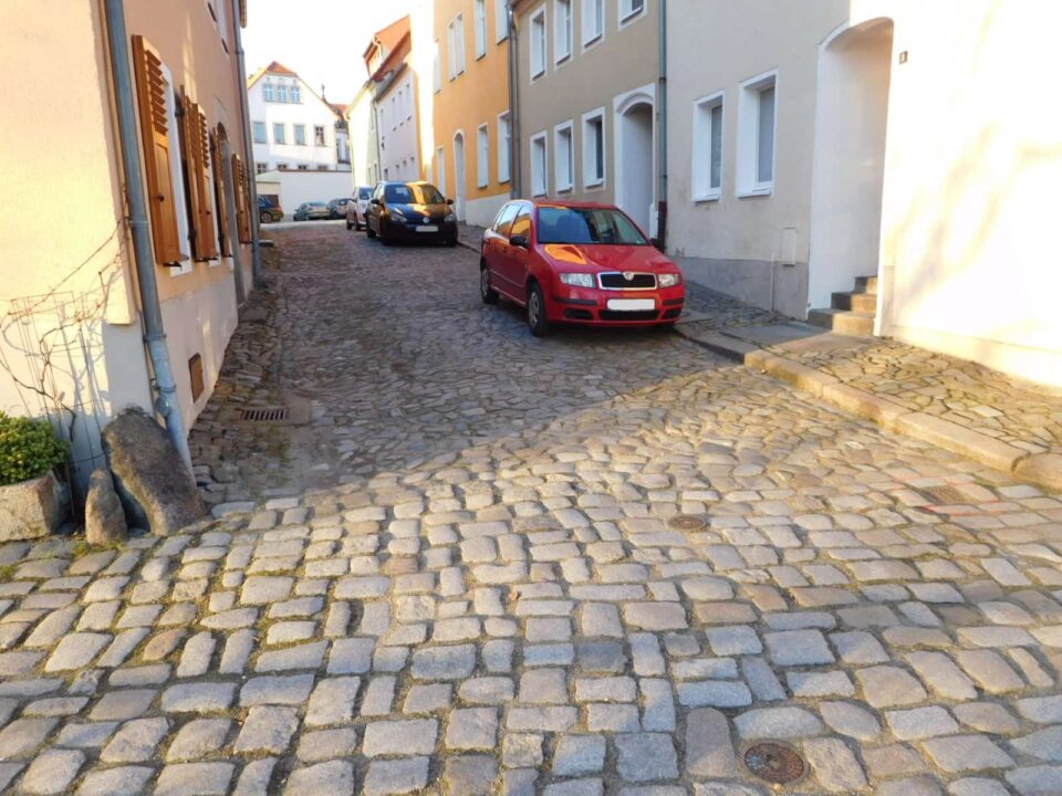 Gepflasterte Straße in einer Altstadt mit geparkten Autos entlang der Häuserfassaden: Straßensanierung mit ProVI