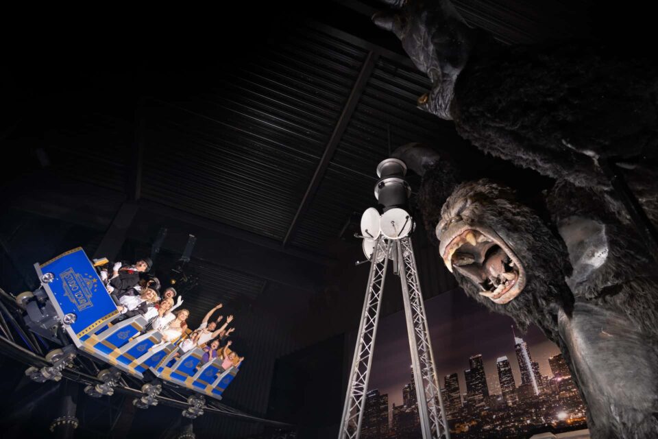 Achterbahnwagen mit jubelnden Menschen fährt durch eine Indoor-Attraktion, die eine riesige King-Kong-Figur mit bedrohlichem Gesichtsausdruck zeigt. 